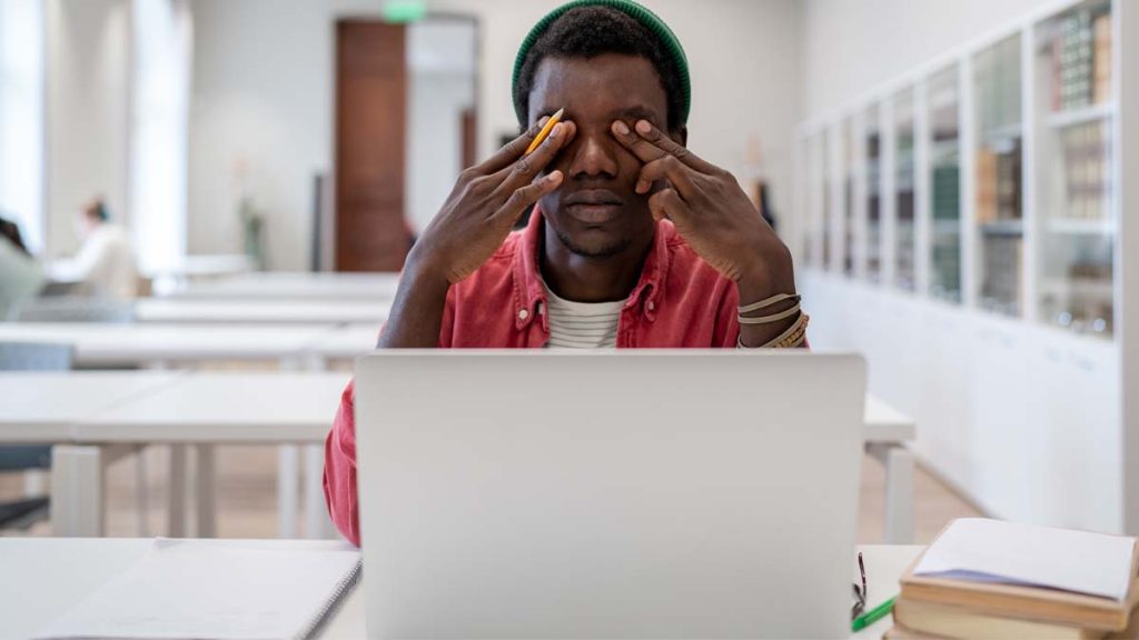 Photo of a man behind a laptop rubbing his eyes